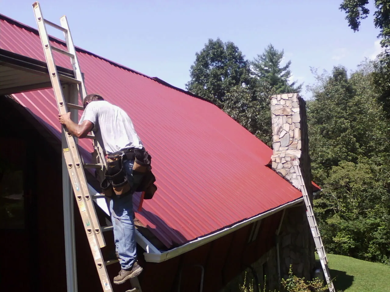 Roofer on a ladder beside a red metal roof and stone chimney — Soffit & Fascia Repair by Roofing Brothers in Plantersville